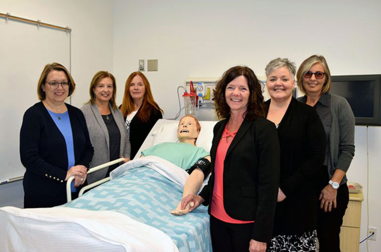 Six School of Nursing administrators and instructors standing around a simulation bed with a mannequin lying on it